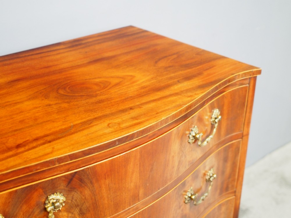 A Hepplewhite style mahogany chest of drawers featuring a serpentine front, four graduated drawers with cast brass handles, boxwood string inlay, and splayed bracket feet. View V
