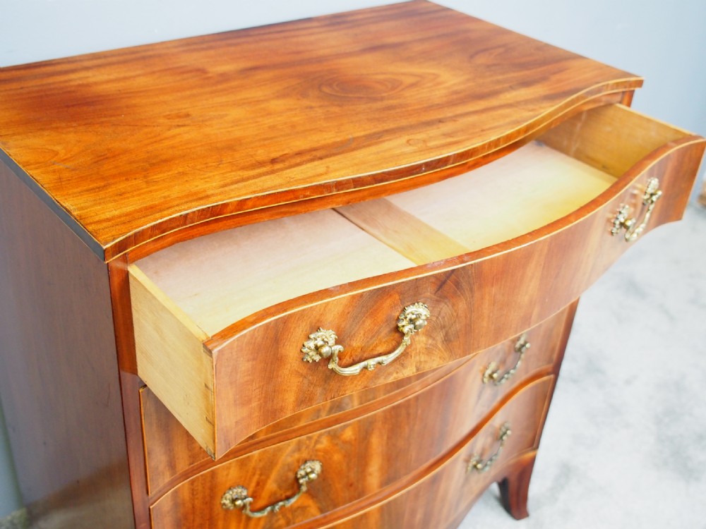 A Hepplewhite style mahogany chest of drawers featuring a serpentine front, four graduated drawers with cast brass handles, boxwood string inlay, and splayed bracket feet. View IX