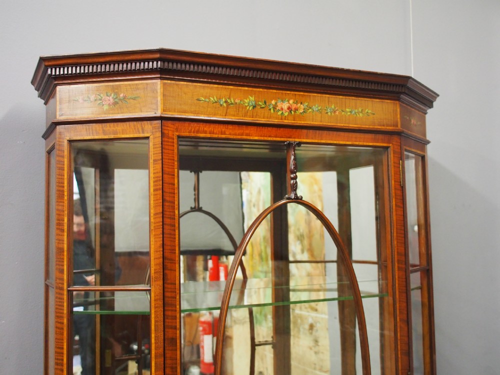 A Sheraton style bowfront display cabinet crafted from warm wood with floral painted swags, oval astragal glazed doors, and glass shelving. View IV