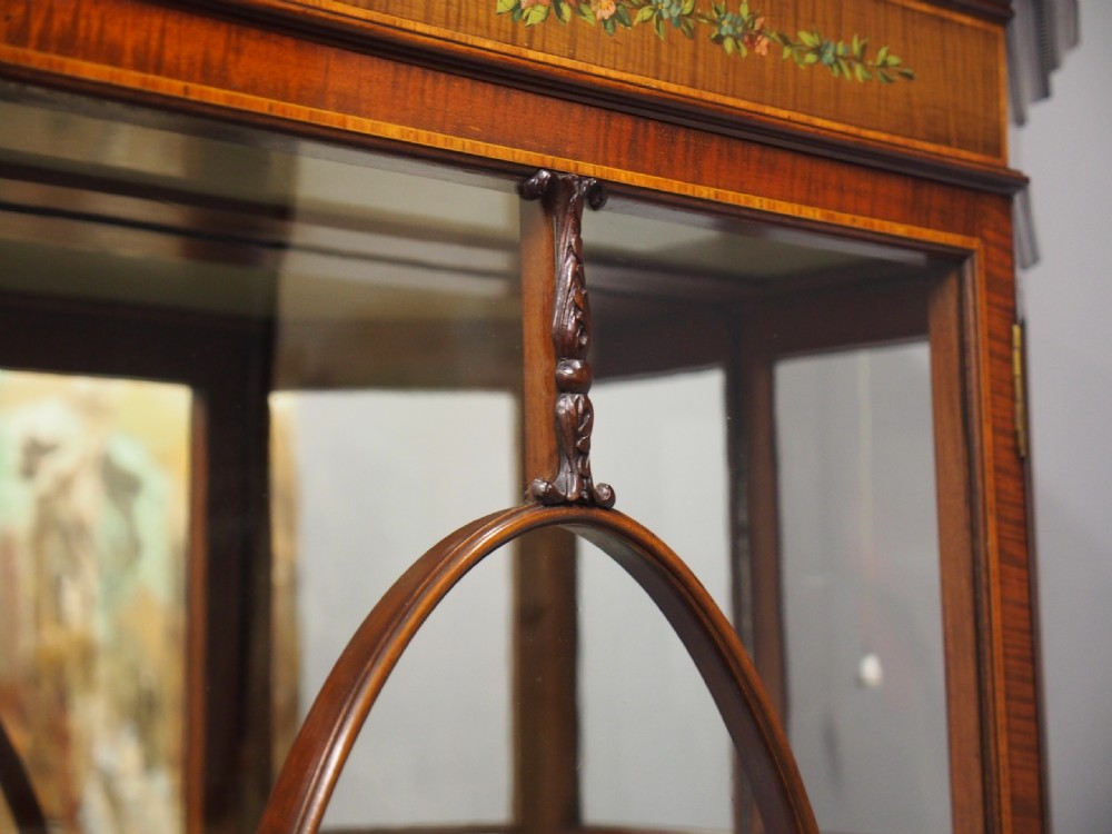 A Sheraton style bowfront display cabinet crafted from warm wood with floral painted swags, oval astragal glazed doors, and glass shelving. View VI