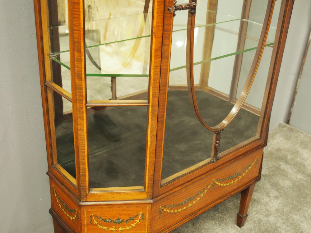 A Sheraton style bowfront display cabinet crafted from warm wood with floral painted swags, oval astragal glazed doors, and glass shelving. View VIII