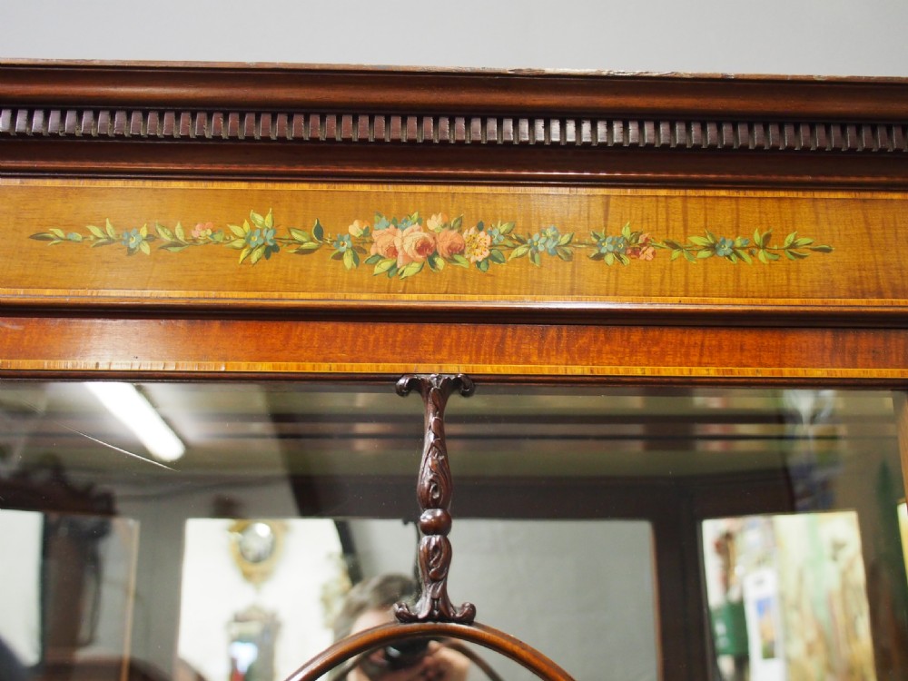 A Sheraton style bowfront display cabinet crafted from warm wood with floral painted swags, oval astragal glazed doors, and glass shelving. View XI