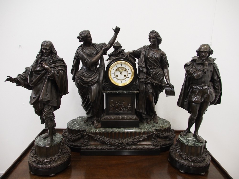 A three-piece French antique clock garniture featuring a central mantel clock flanked by classical female figures and two standalone bronze male sculptures on dark marble bases. View XII