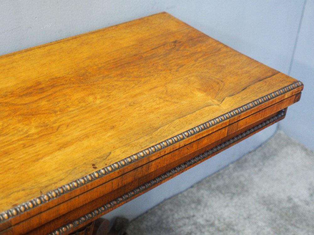 A George III rosewood games table featuring a beaded fold-over top, central turned pedestal support, and four splayed legs with brass castors. View IV