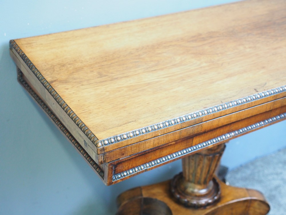 A George III rosewood games table featuring a beaded fold-over top, central turned pedestal support, and four splayed legs with brass castors. View V