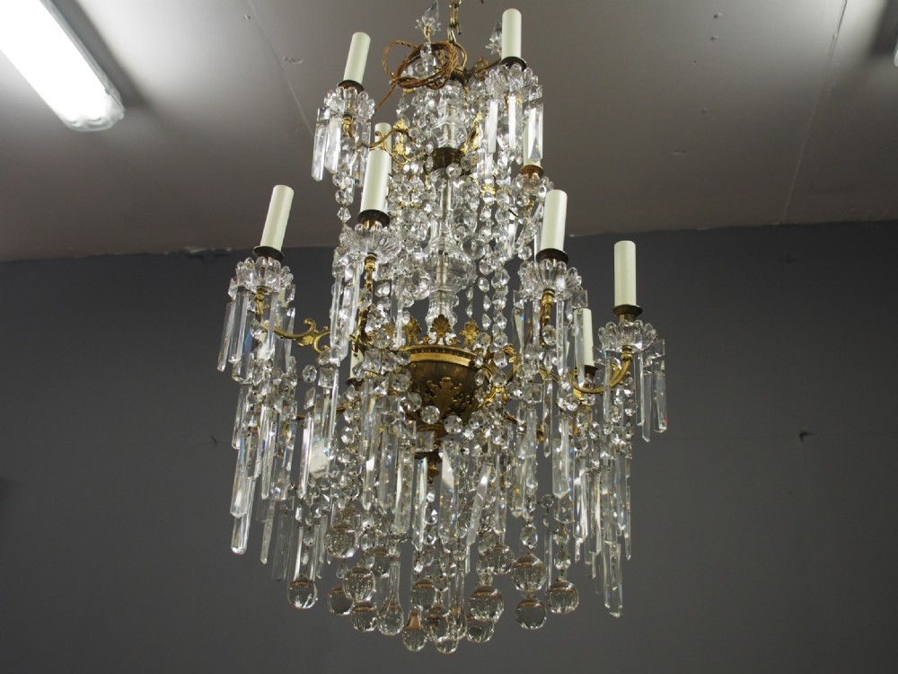A two-tiered ceiling chandelier featuring a gilded brass frame adorned with multiple hanging glass prisms and six candle-style light sockets. View II