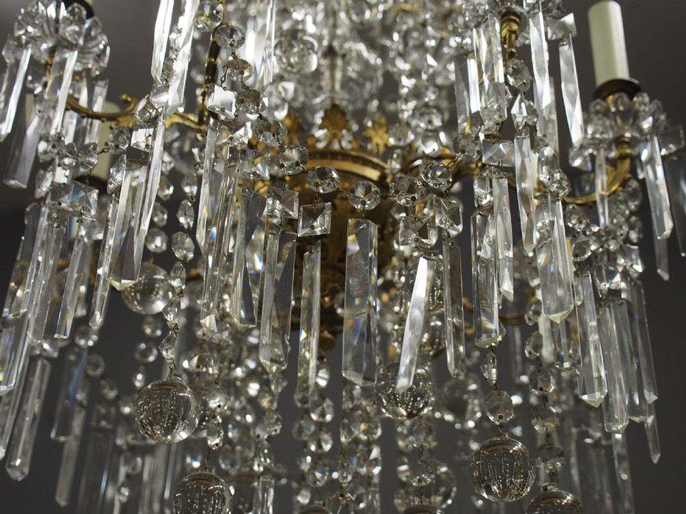 A two-tiered ceiling chandelier featuring a gilded brass frame adorned with multiple hanging glass prisms and six candle-style light sockets. View III