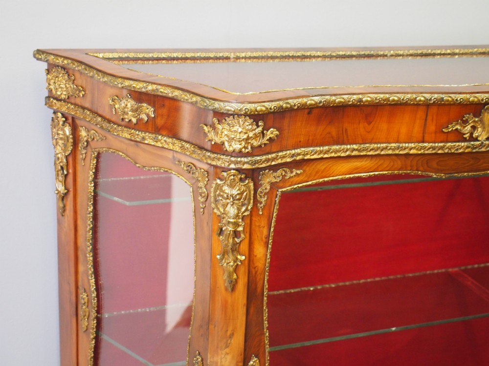 A Victorian yew wood vitrine display cabinet with gilt-bronze ormolu mounts, curved glass front, and red velvet lining. View VI