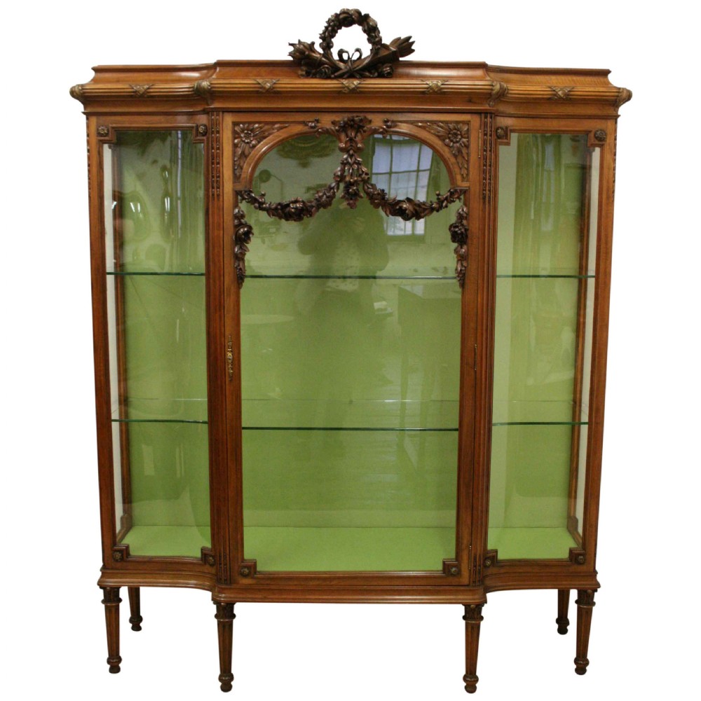 A carved walnut French display cabinet featuring glazed doors, glass shelves, green fabric interior lining, and turned fluted legs.