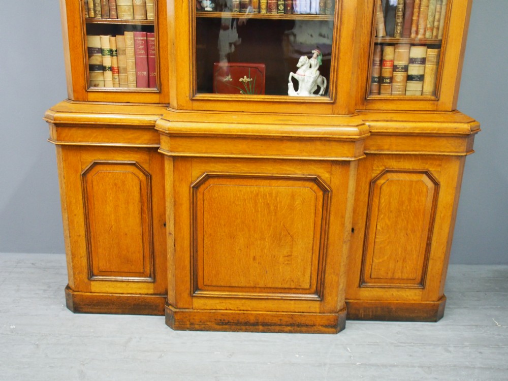 A Victorian golden oak breakfront bookcase featuring three arch-glazed upper doors above three solid timber panel cupboard doors. View IV