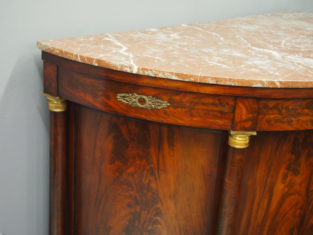 A French Empire demilune mahogany side cabinet featuring a reddish-pink variegated marble top, column flanks with gilt accents, and two central cupboard doors. View VI