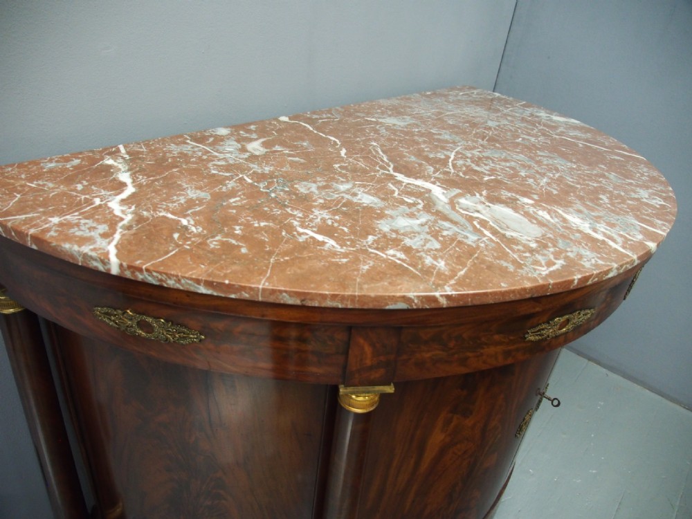 A French Empire demilune mahogany side cabinet featuring a reddish-pink variegated marble top, column flanks with gilt accents, and two central cupboard doors. View VII