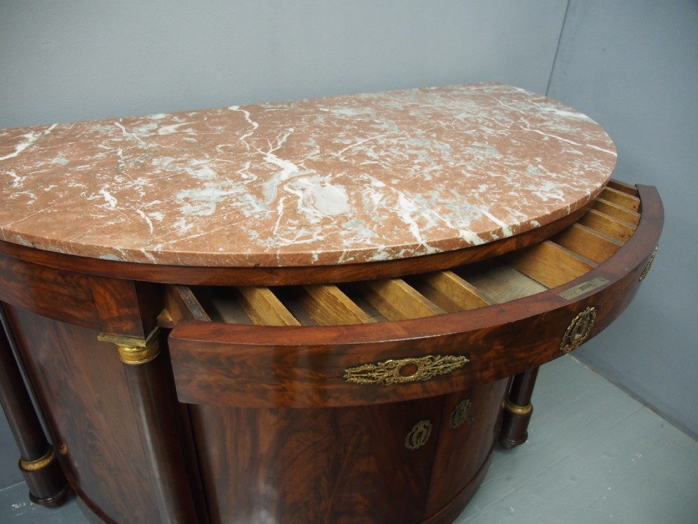 A French Empire demilune mahogany side cabinet featuring a reddish-pink variegated marble top, column flanks with gilt accents, and two central cupboard doors. View VIII