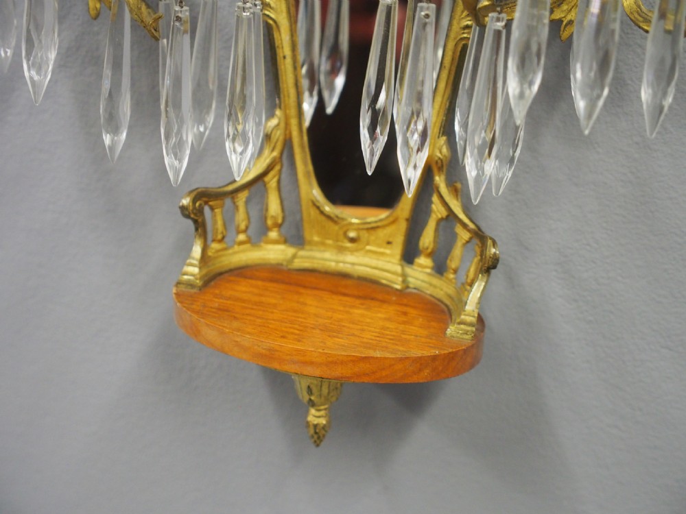 A pair of ornate French Neoclassical ormolu wall sconces featuring oval mirrored backs, multi-arm candleholders, cut crystal prisms, and top trophy crests. View IX