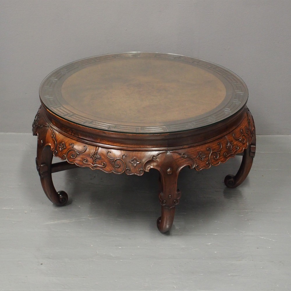A circular low Chinese hardwood table featuring a glass top over a fretwork border, carved aprons, and curved legs.