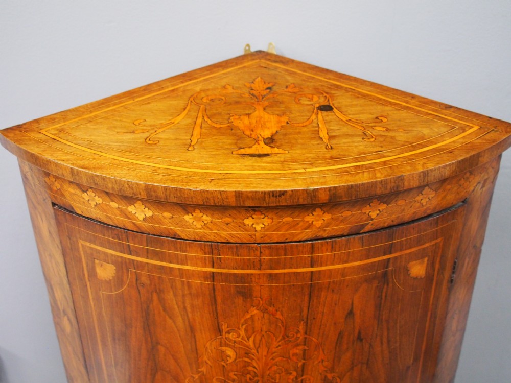 A floor-standing Dutch walnut corner cabinet featuring a curved front door embellished with figurative and floral marquetry inlays resting on carved legs. View VI
