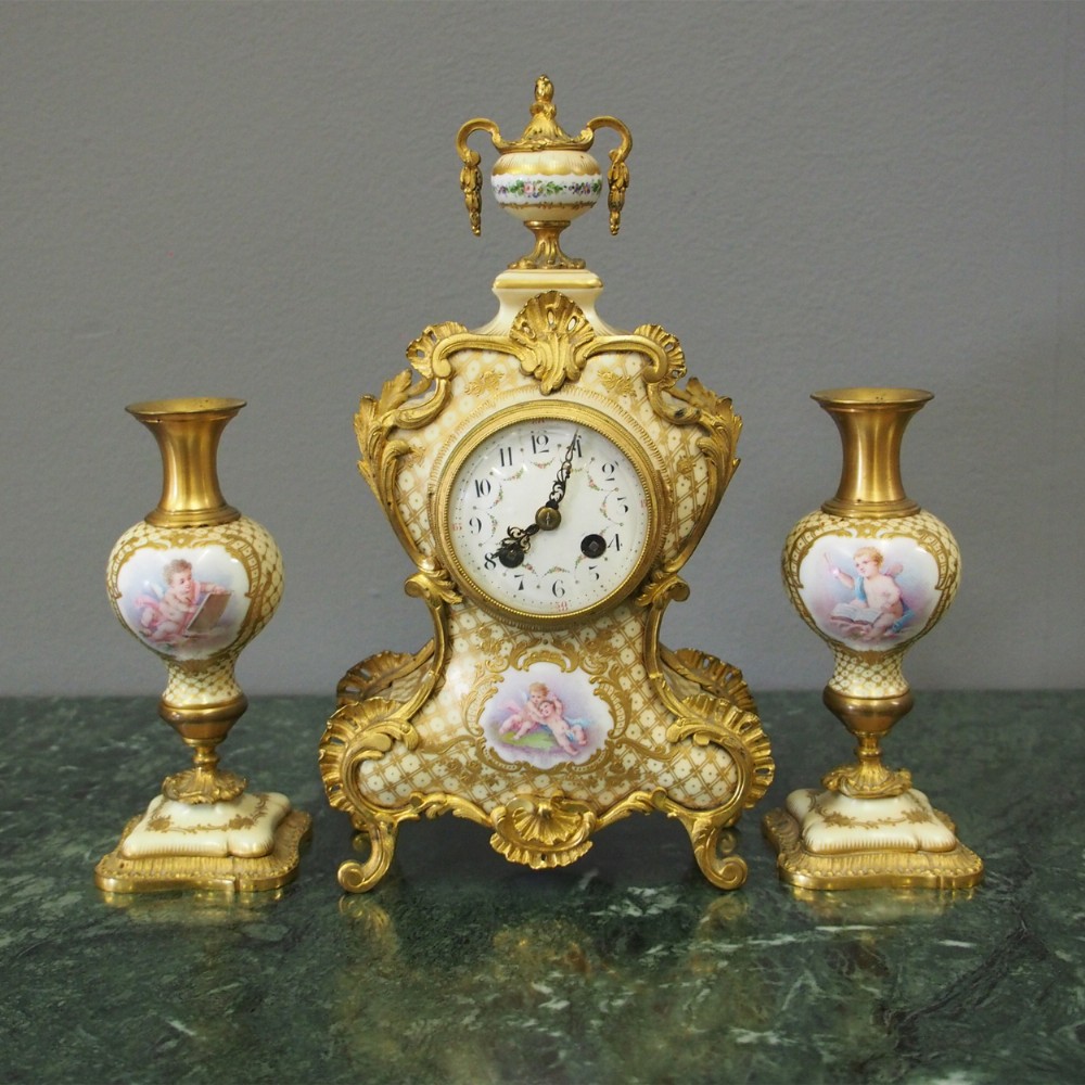 A three-piece French porcelain clock garniture set featuring a central rococo-style clock flanked by two matching vases decorated with hand-painted cherubs and gilded mounts.