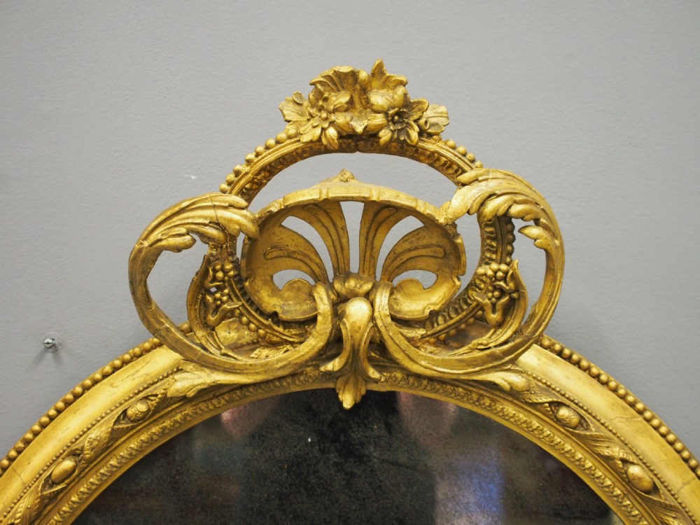 A Victorian oval giltwood wall mirror featuring an intricate carved cresting at the top, scrollwork detail at the bottom, beaded trim, and two small display shelves on the sides. View III