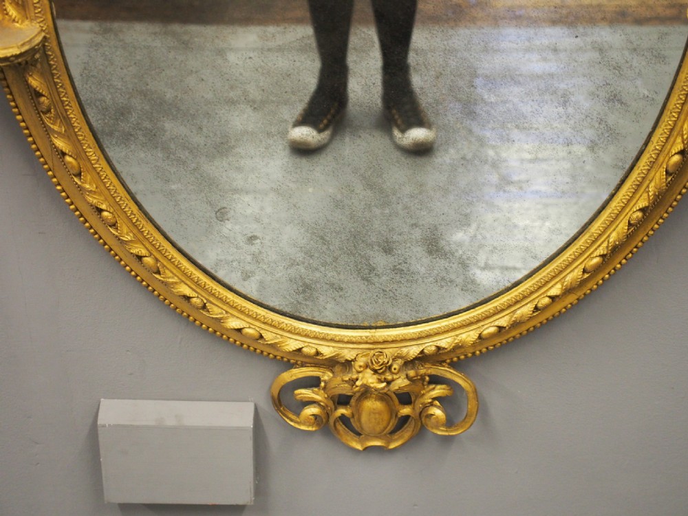 A Victorian oval giltwood wall mirror featuring an intricate carved cresting at the top, scrollwork detail at the bottom, beaded trim, and two small display shelves on the sides. View VI