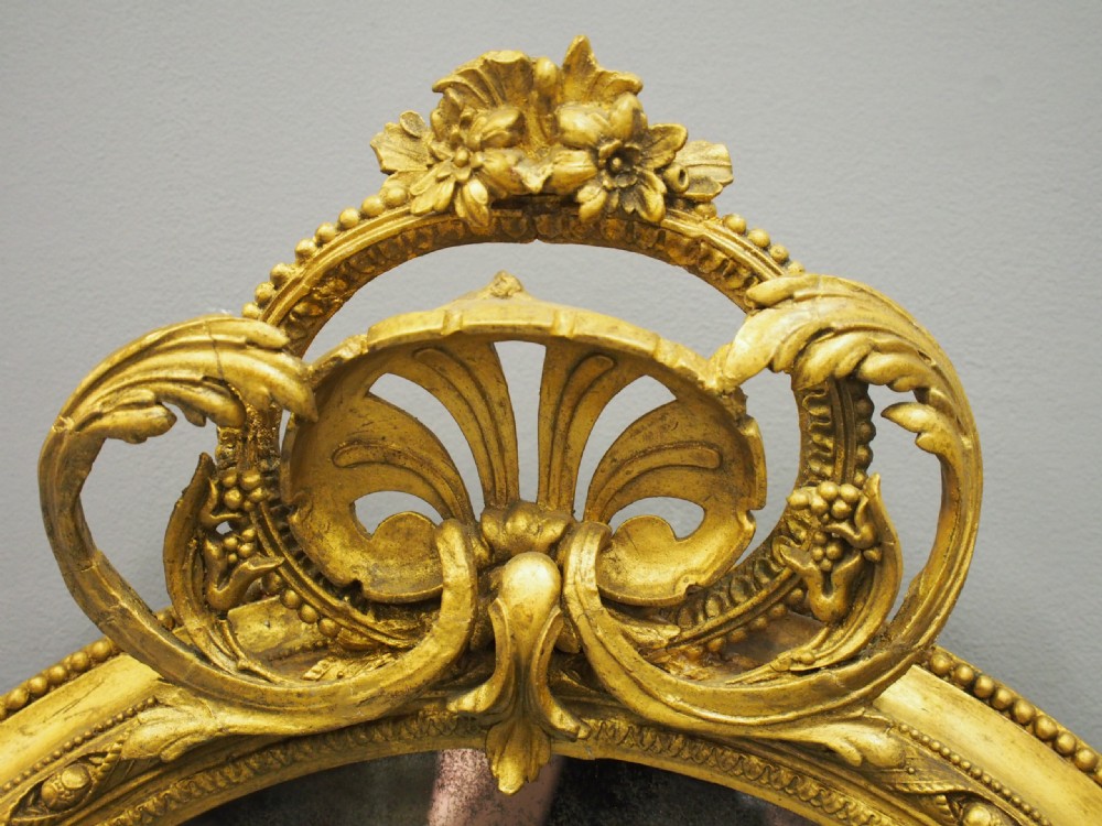 A Victorian oval giltwood wall mirror featuring an intricate carved cresting at the top, scrollwork detail at the bottom, beaded trim, and two small display shelves on the sides. View VII