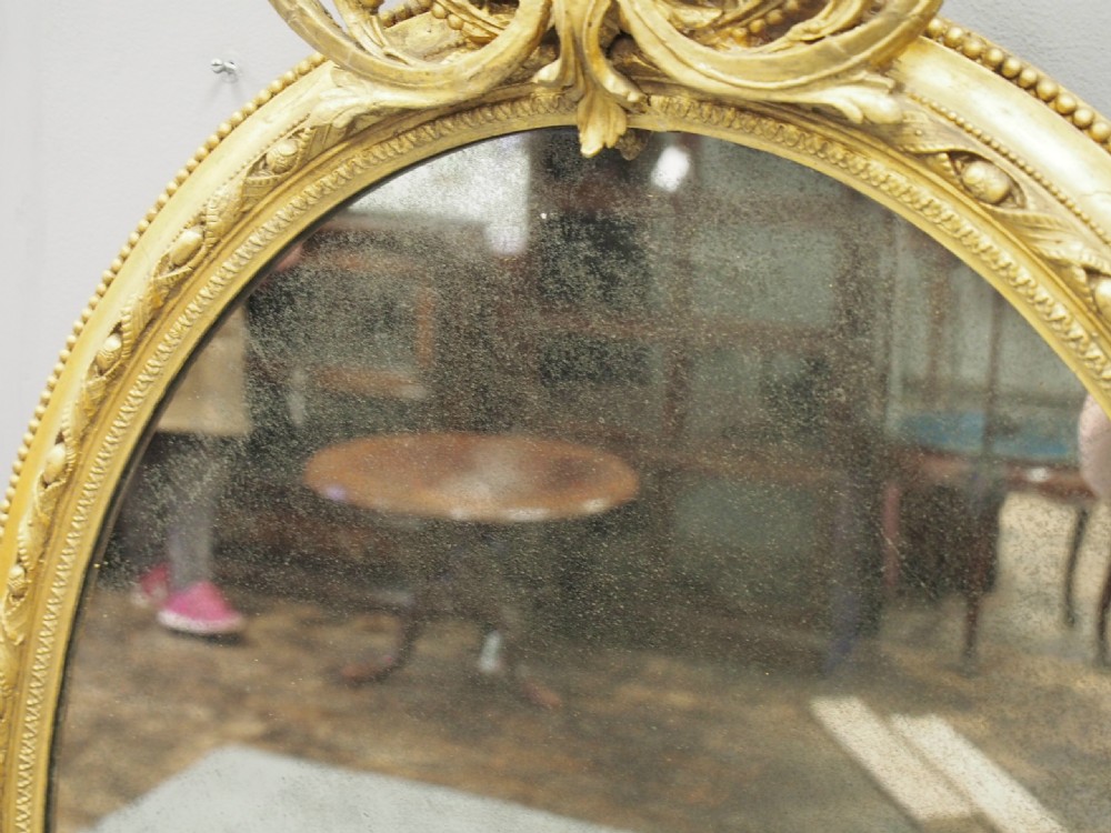 A Victorian oval giltwood wall mirror featuring an intricate carved cresting at the top, scrollwork detail at the bottom, beaded trim, and two small display shelves on the sides. View VIII