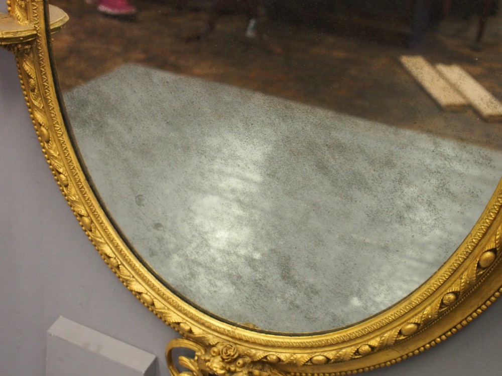 A Victorian oval giltwood wall mirror featuring an intricate carved cresting at the top, scrollwork detail at the bottom, beaded trim, and two small display shelves on the sides. View IX