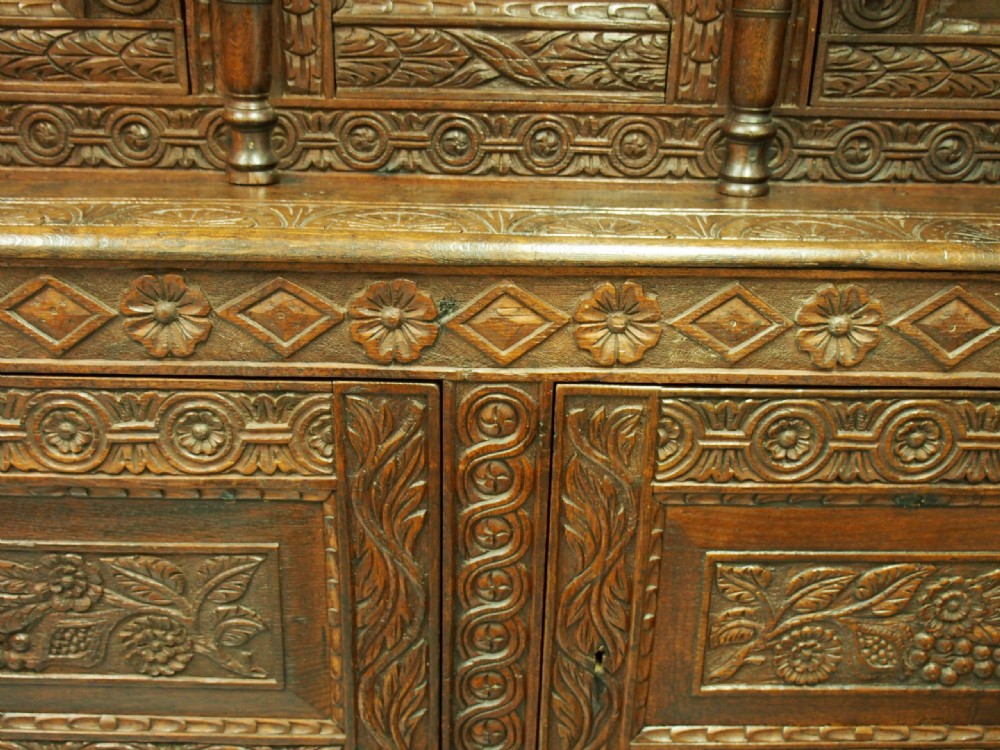 A dark carved oak Charles II court cupboard featuring intricate floral relief panels, turned supports, inscribed initials, and decorative brass hinges. View VII