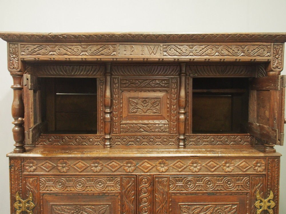 A dark carved oak Charles II court cupboard featuring intricate floral relief panels, turned supports, inscribed initials, and decorative brass hinges. View XII