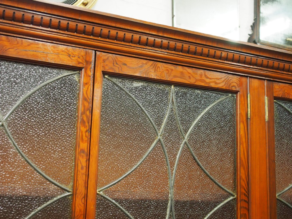 A large Victorian pitch pine bookcase cabinet featuring four upper glazed doors with geometric leaded glass and four lower panelled cupboard doors. View III