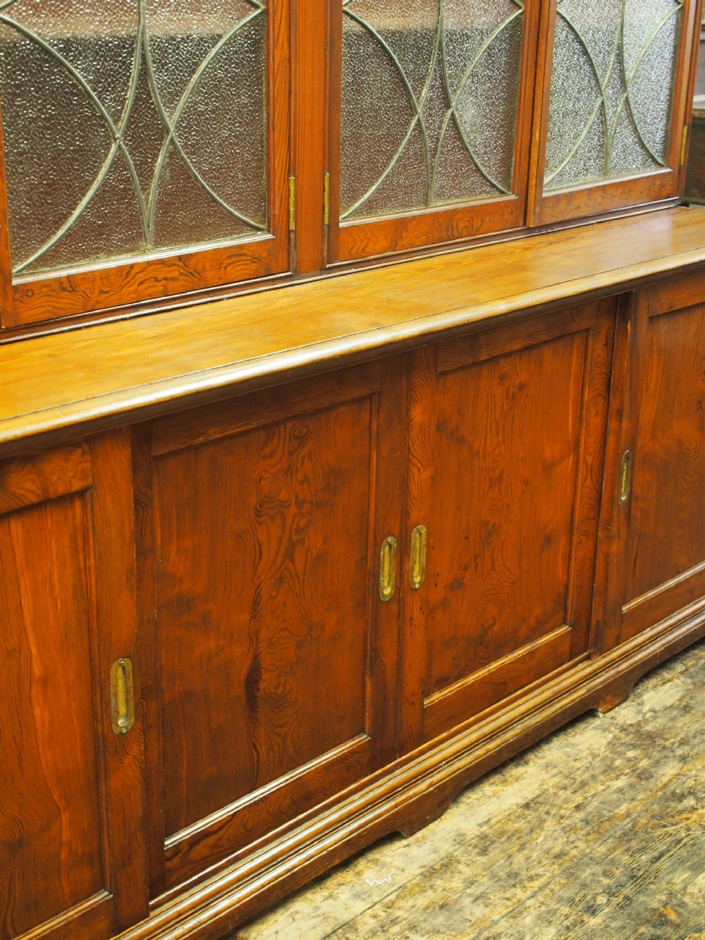 A large Victorian pitch pine bookcase cabinet featuring four upper glazed doors with geometric leaded glass and four lower panelled cupboard doors. View VII