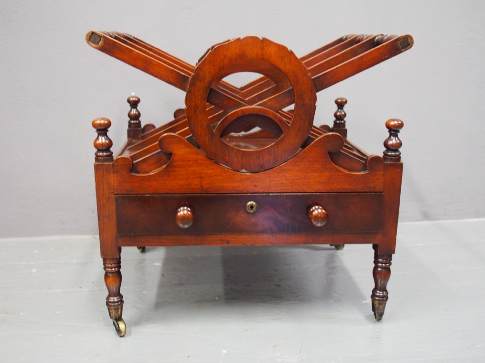 A George IV mahogany Canterbury featuring carved circular dividers, turned corner posts, a single base drawer with two wooden turned knobs, and legs set on brass castors. View II