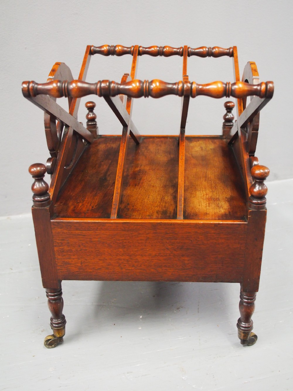 A George IV mahogany Canterbury featuring carved circular dividers, turned corner posts, a single base drawer with two wooden turned knobs, and legs set on brass castors. View V