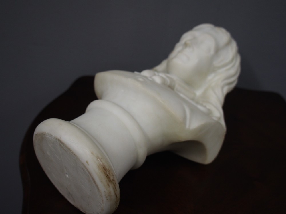 A white marble carved portrait bust depicting a gentleman with long flowing hair and period attire mounted on a round socle base. View VI