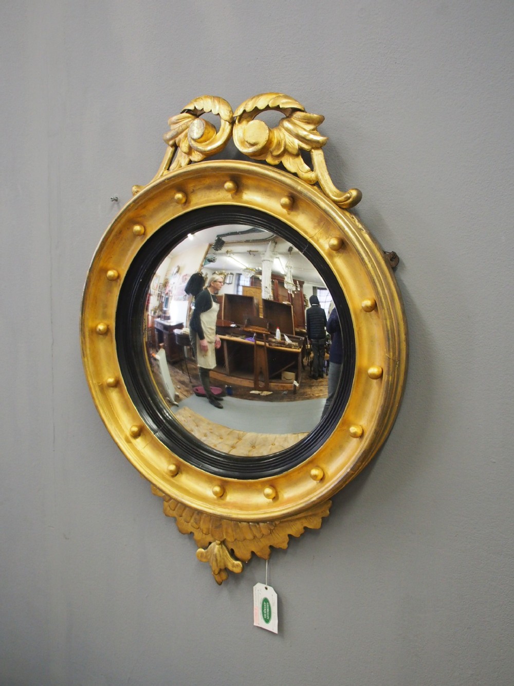 A circular gold giltwood convex mirror featuring a carved acanthus leaf crest, ebonised inner slip, and spherule studs around the frame. View II