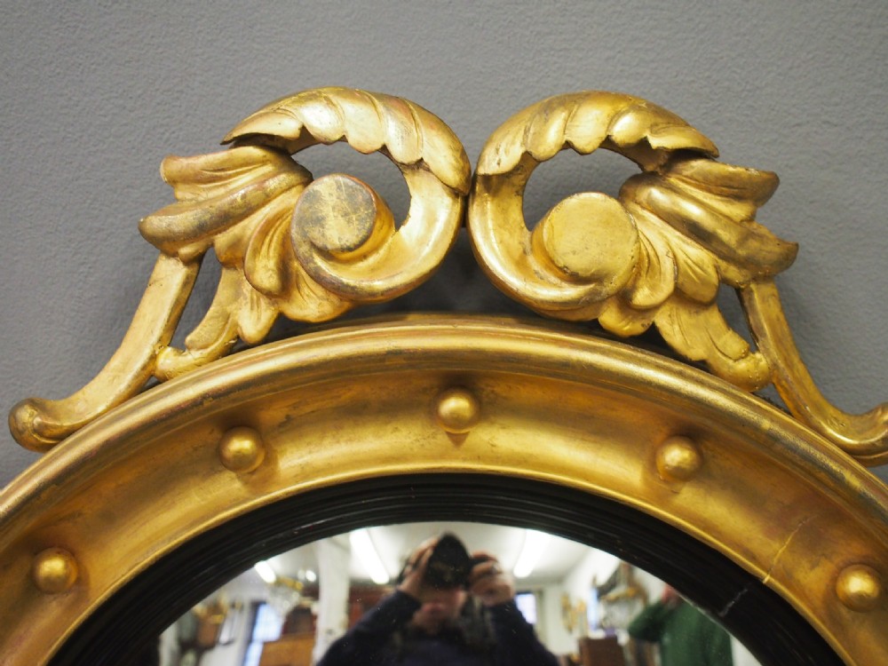 A circular gold giltwood convex mirror featuring a carved acanthus leaf crest, ebonised inner slip, and spherule studs around the frame. View III