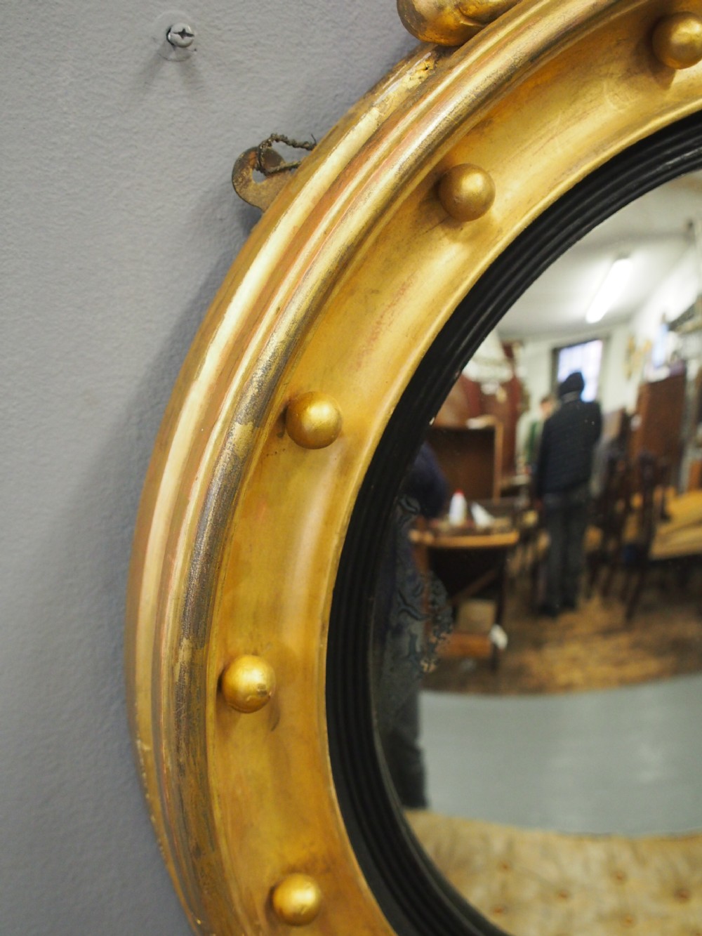 A circular gold giltwood convex mirror featuring a carved acanthus leaf crest, ebonised inner slip, and spherule studs around the frame. View IV