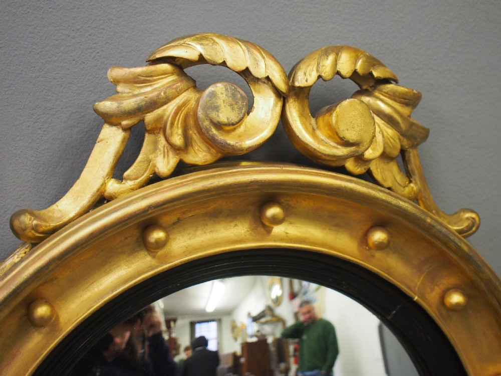 A circular gold giltwood convex mirror featuring a carved acanthus leaf crest, ebonised inner slip, and spherule studs around the frame. View VI