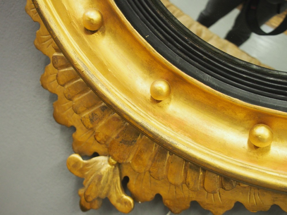 A circular gold giltwood convex mirror featuring a carved acanthus leaf crest, ebonised inner slip, and spherule studs around the frame. View IX