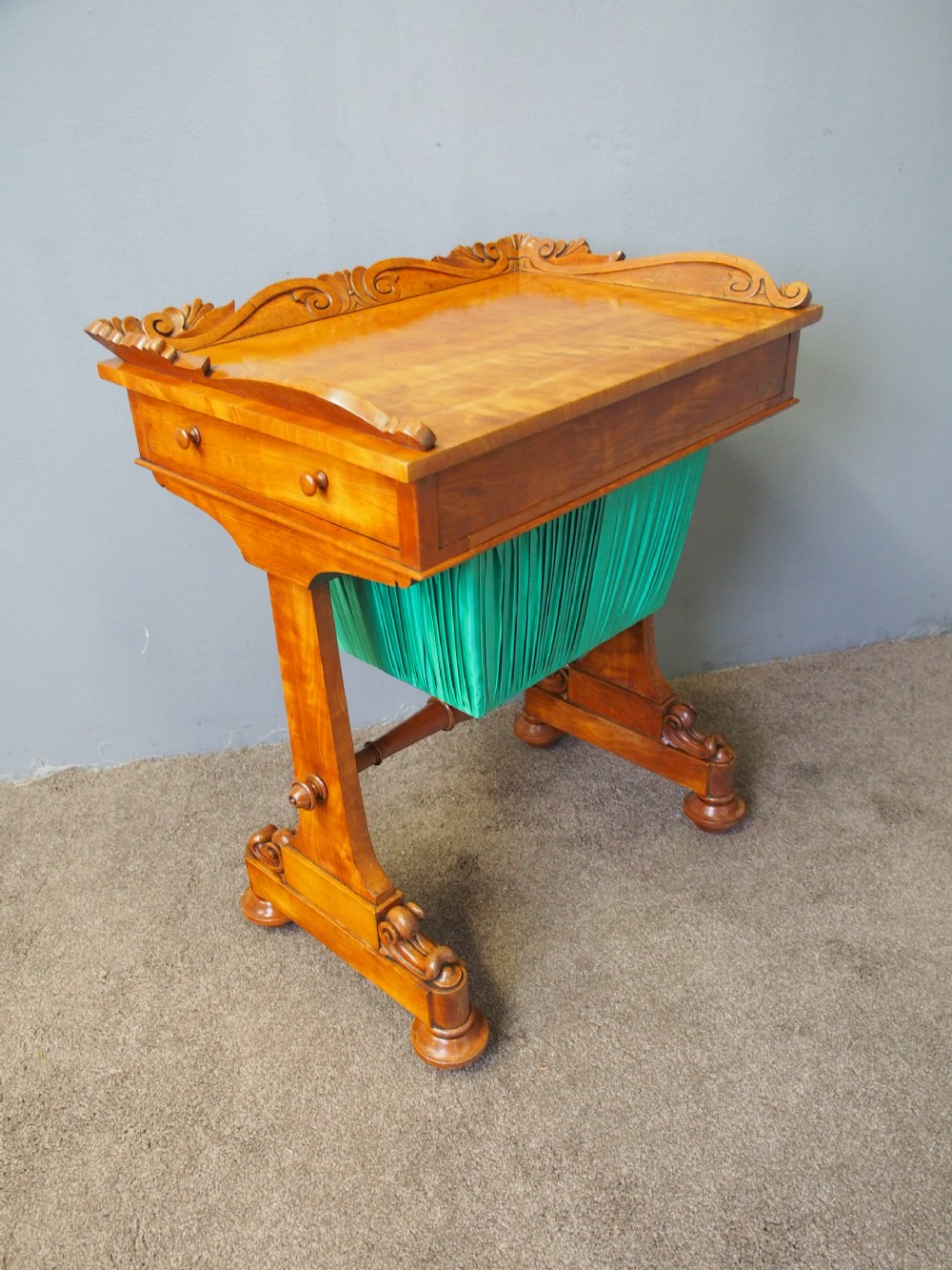 A Victorian satinwood work table featuring a carved top gallery, single drawer, pleated green silk work well, and carved trestle legs. View II