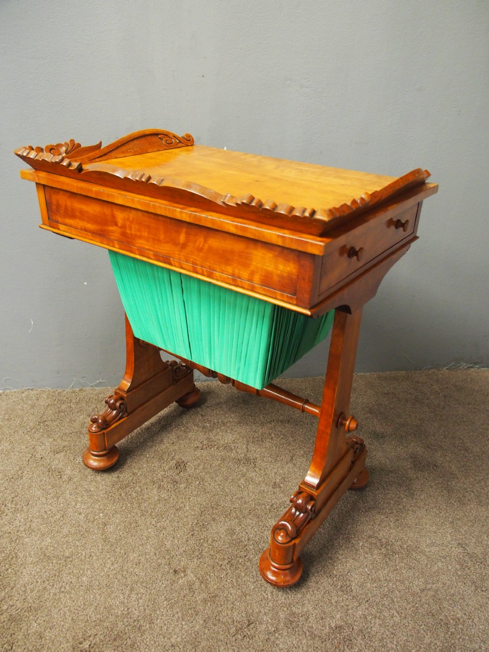 A Victorian satinwood work table featuring a carved top gallery, single drawer, pleated green silk work well, and carved trestle legs. View IX