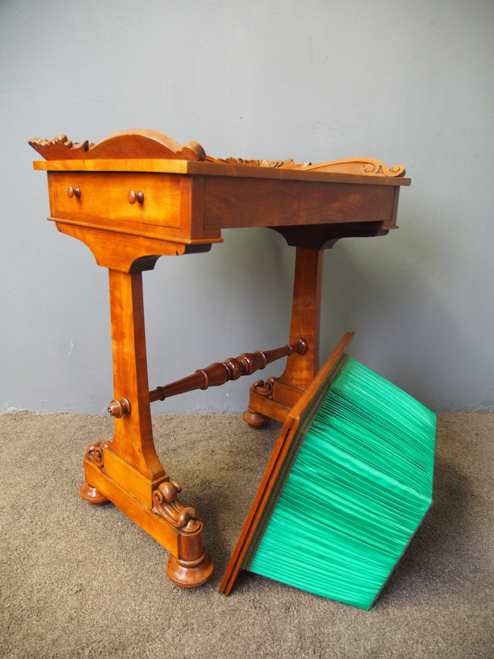 A Victorian satinwood work table featuring a carved top gallery, single drawer, pleated green silk work well, and carved trestle legs. View XIV