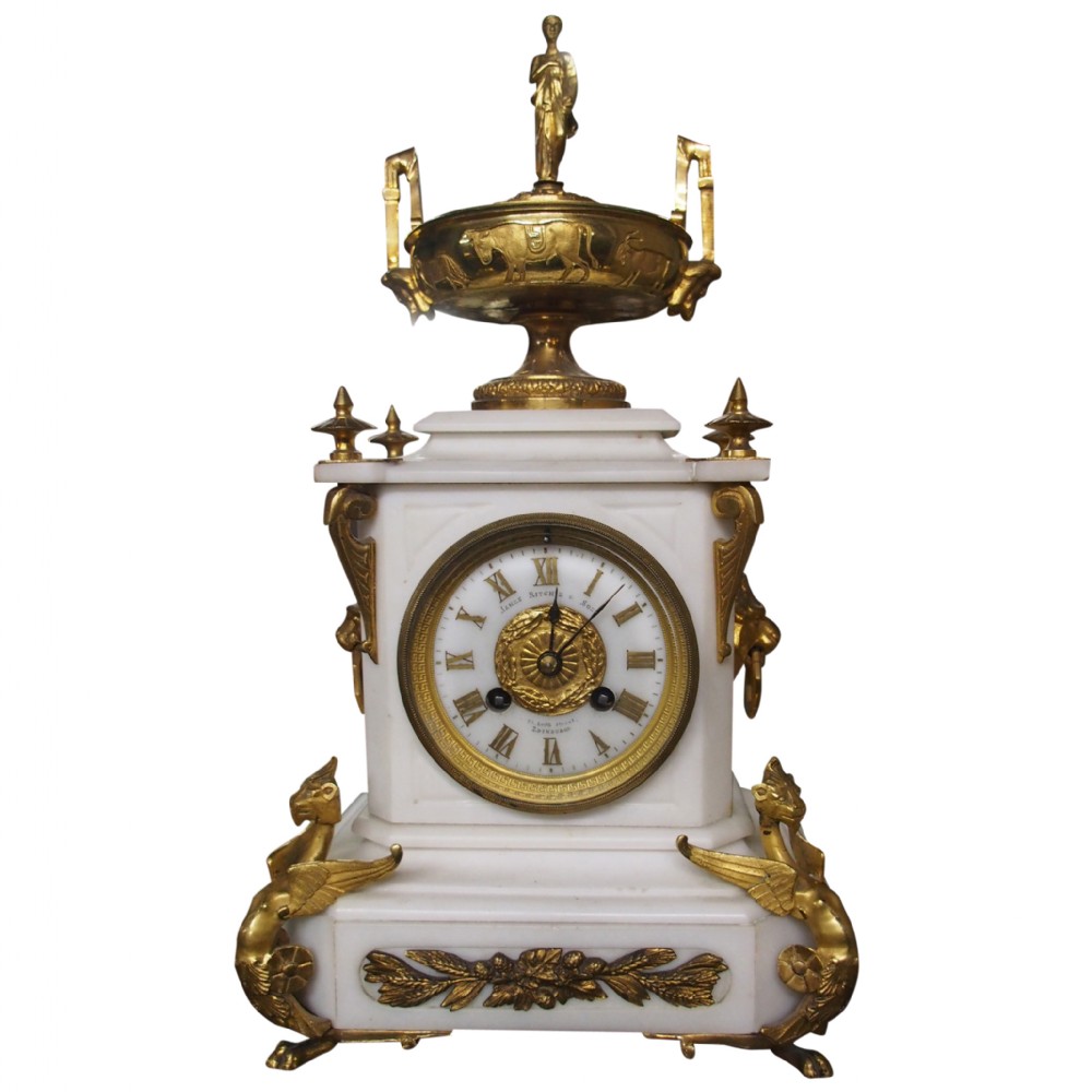 A white marble mantel clock featuring gilt bronze mounts, winged griffin corner supports, and a classical urn finial topped with a standing figure.