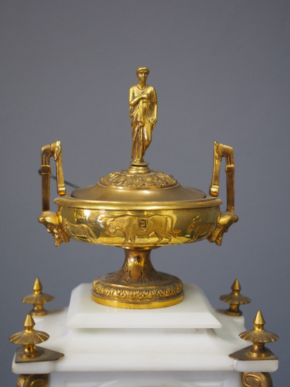 A white marble mantel clock featuring gilt bronze mounts, winged griffin corner supports, and a classical urn finial topped with a standing figure. View V