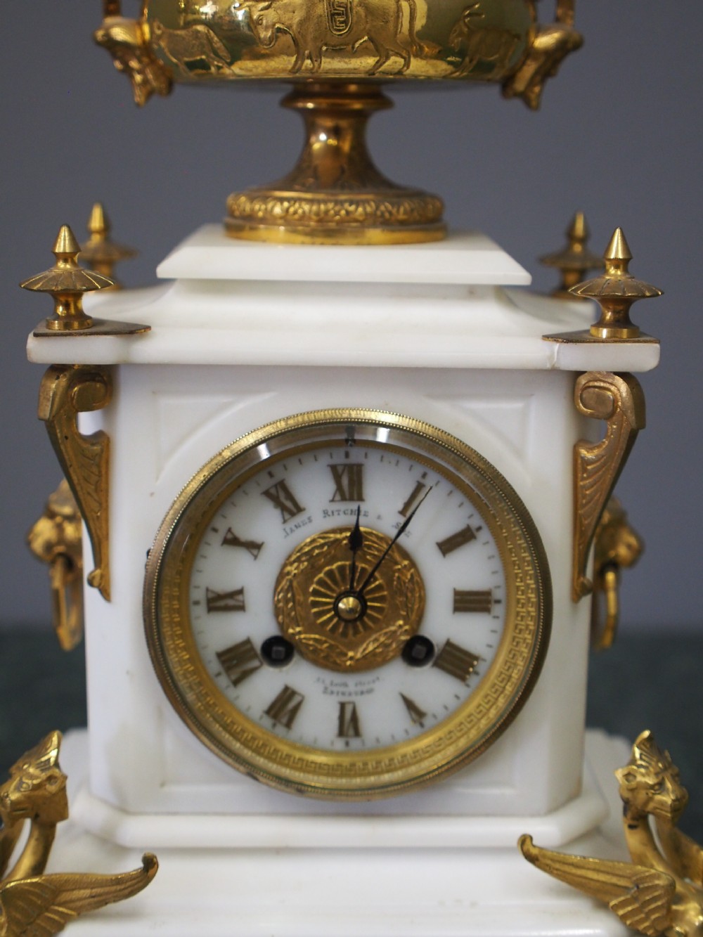 A white marble mantel clock featuring gilt bronze mounts, winged griffin corner supports, and a classical urn finial topped with a standing figure. View VI