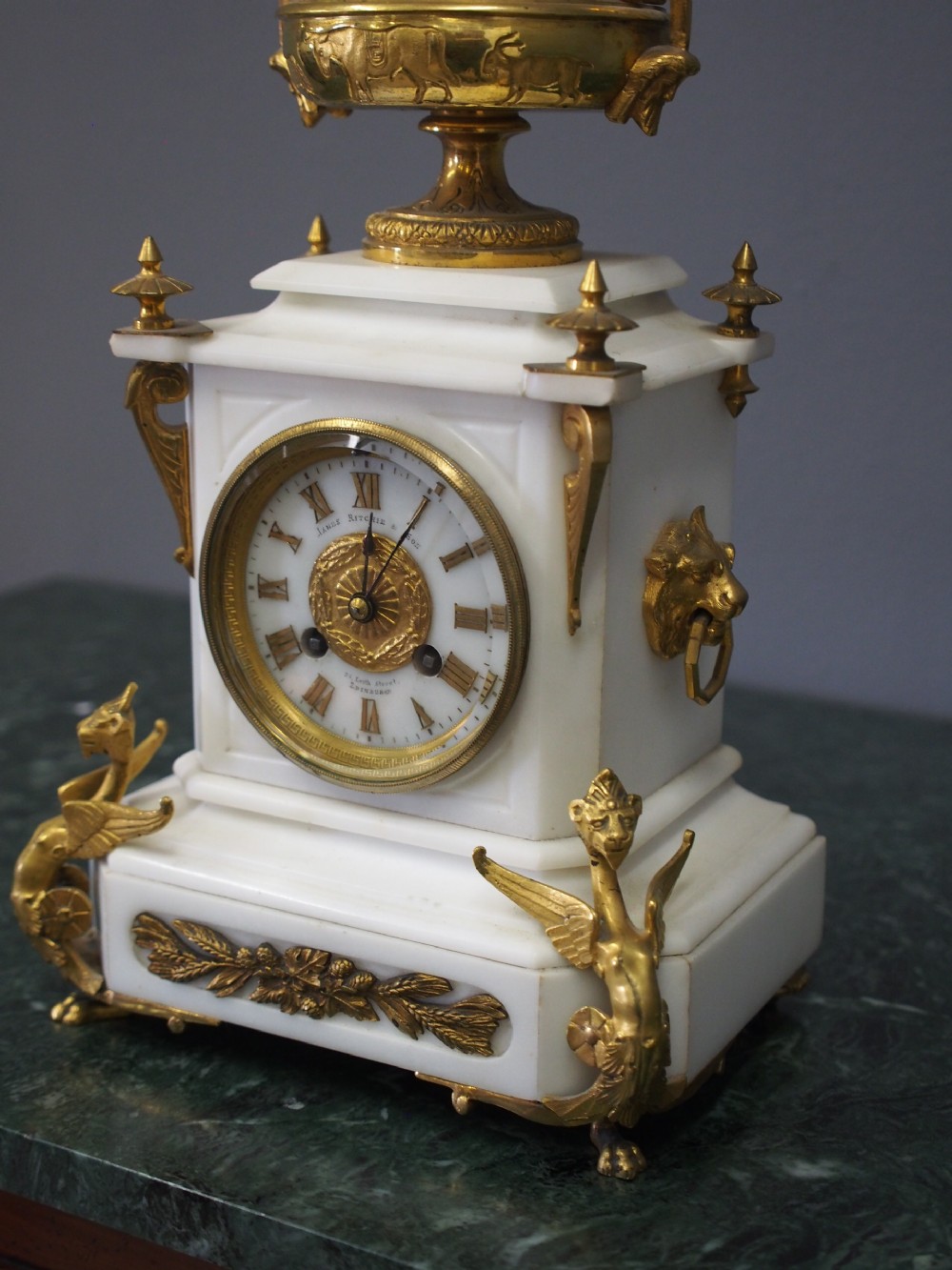 A white marble mantel clock featuring gilt bronze mounts, winged griffin corner supports, and a classical urn finial topped with a standing figure. View VII