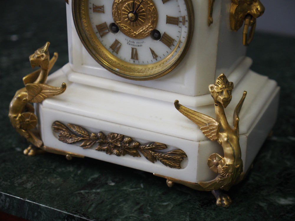 A white marble mantel clock featuring gilt bronze mounts, winged griffin corner supports, and a classical urn finial topped with a standing figure. View VIII