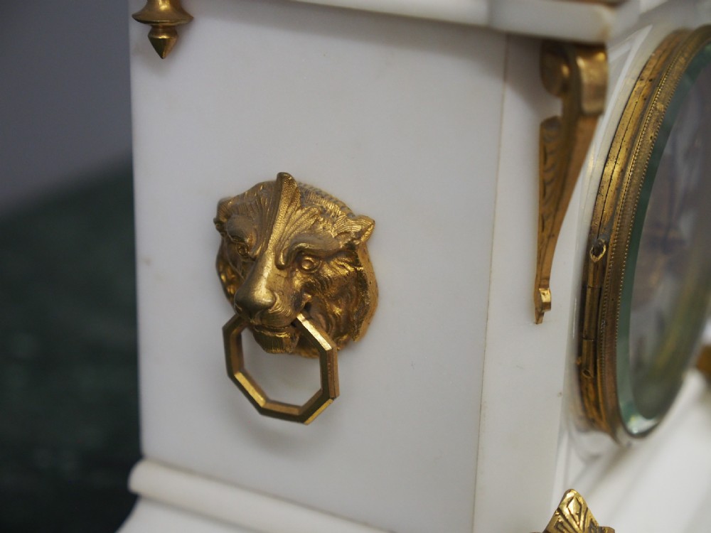 A white marble mantel clock featuring gilt bronze mounts, winged griffin corner supports, and a classical urn finial topped with a standing figure. View IX