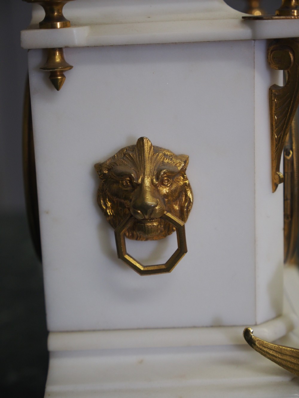 A white marble mantel clock featuring gilt bronze mounts, winged griffin corner supports, and a classical urn finial topped with a standing figure. View X
