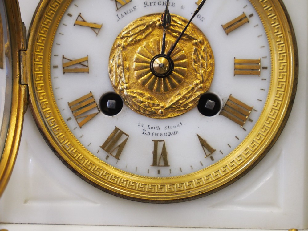 A white marble mantel clock featuring gilt bronze mounts, winged griffin corner supports, and a classical urn finial topped with a standing figure. View XVI