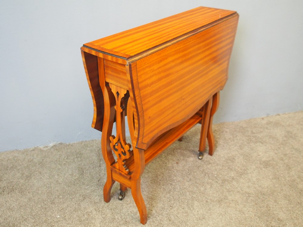 An Edwardian satinwood Sutherland table shown folded with curved drop-leaf sides, fretwork side supports, and brass castors. View II